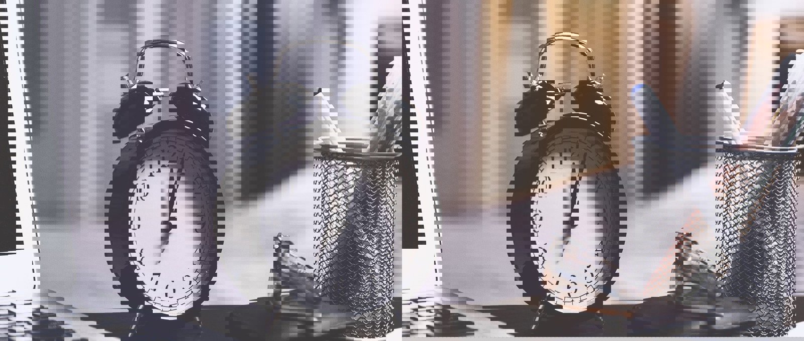 A black analogue clock on a desk beside a pen pot and laptop.