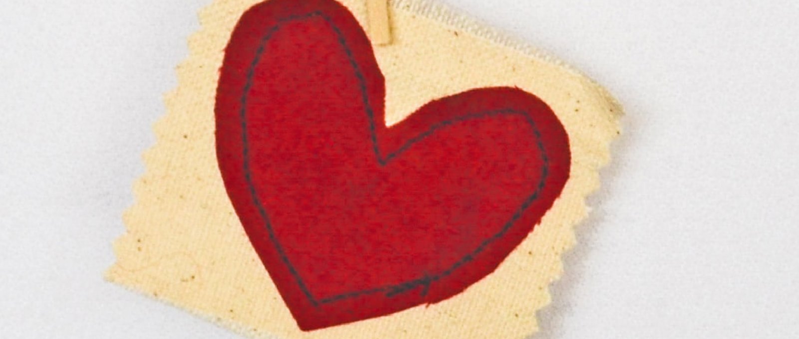 A red heart sewn onto cream fabric, hanging from a piece of string by a peg. 