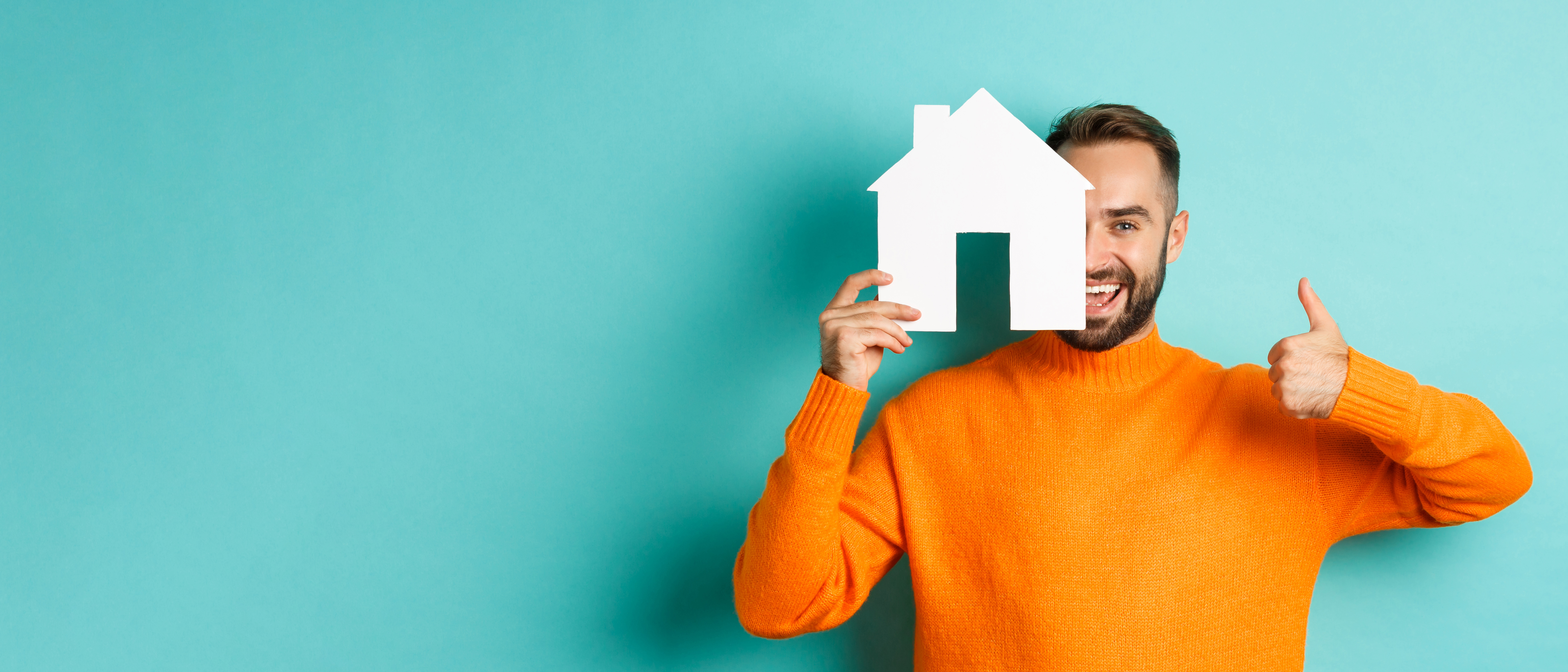 Photo of a smiling man in an orange sweater standing in front of a turquoise background. He is holding a white cut-out house shape half across his face and also giving us a thumbs-up sign