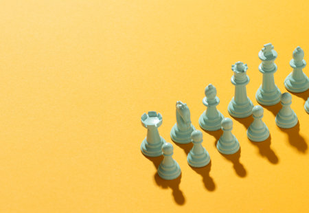 Photograph of a full set of white chess pieces, arranged as they would be for a new game, on a yellow background with strong shadows for each piece