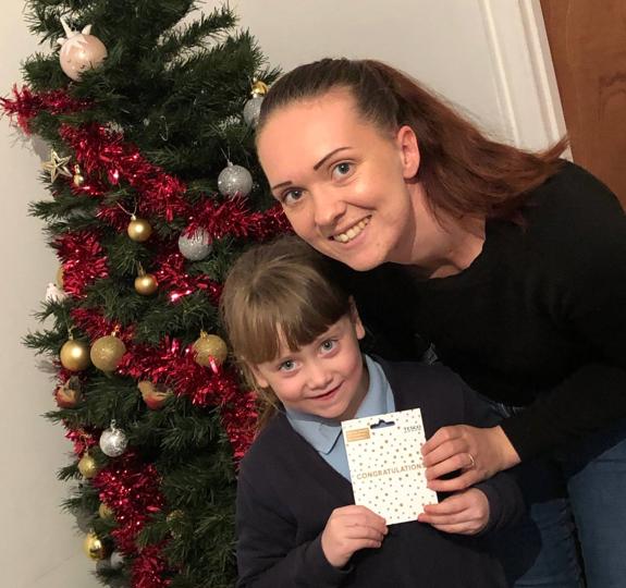 A child and her mother stand in front of a Christmas tree decorated with red tinsel and metallic baubles, holding a shopping voucher.