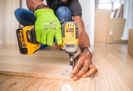 A close up on a person's hands using an electric screwdriver, attaching a metal bracket to a piece of wood.
