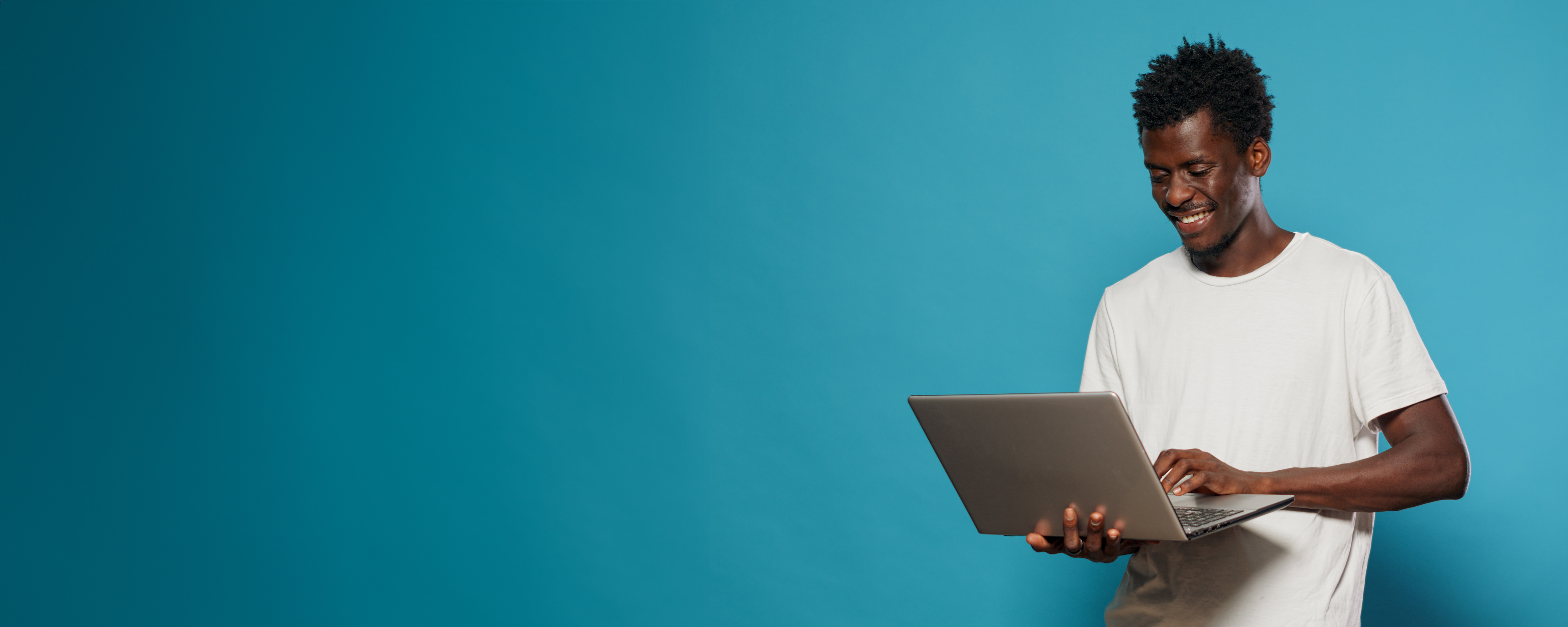 A young man wearing a white T-shirt holding a laptop. He is smiling and standing in front of a blue background.