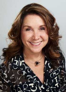 Lindsey smiles to camera in a black and white patterned shirt. She has shoulder length brown hair.