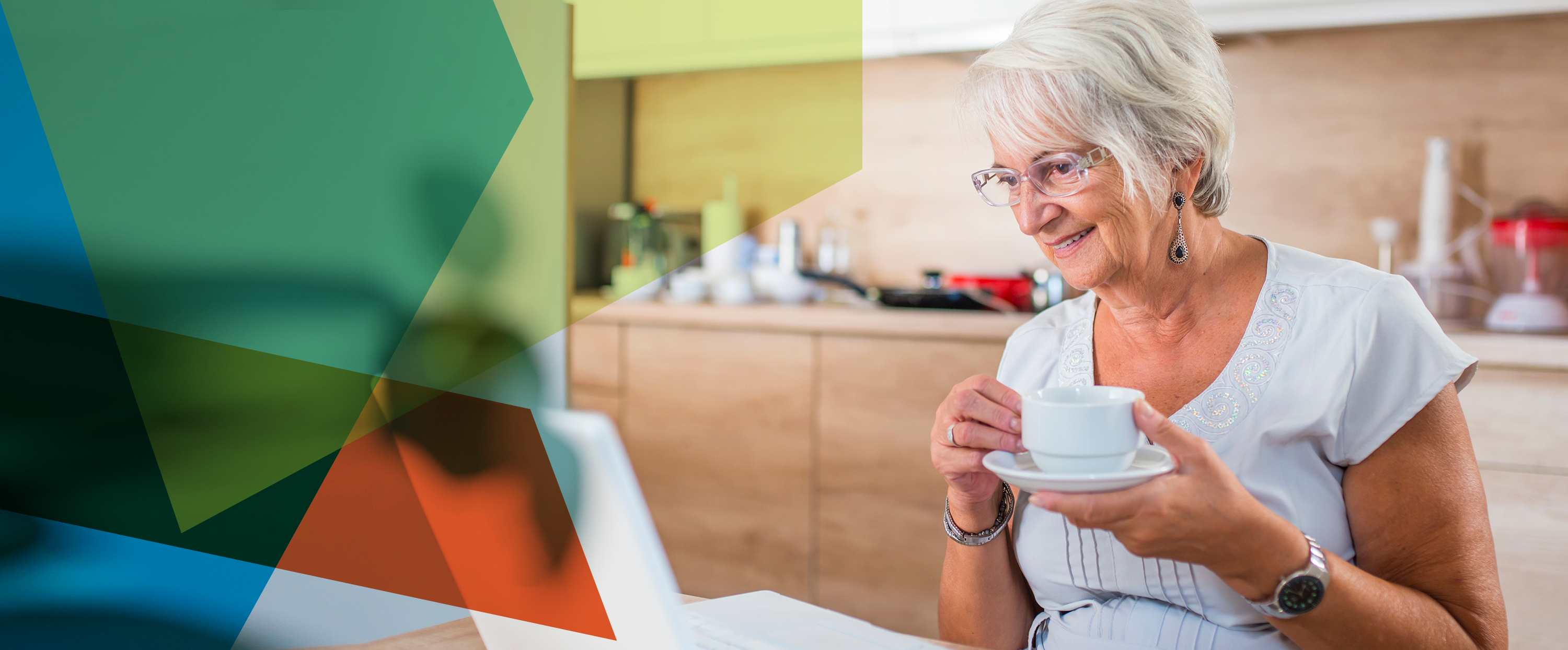 Photograph of an older woman holding a teacup in her kitchen and looking at a laptop