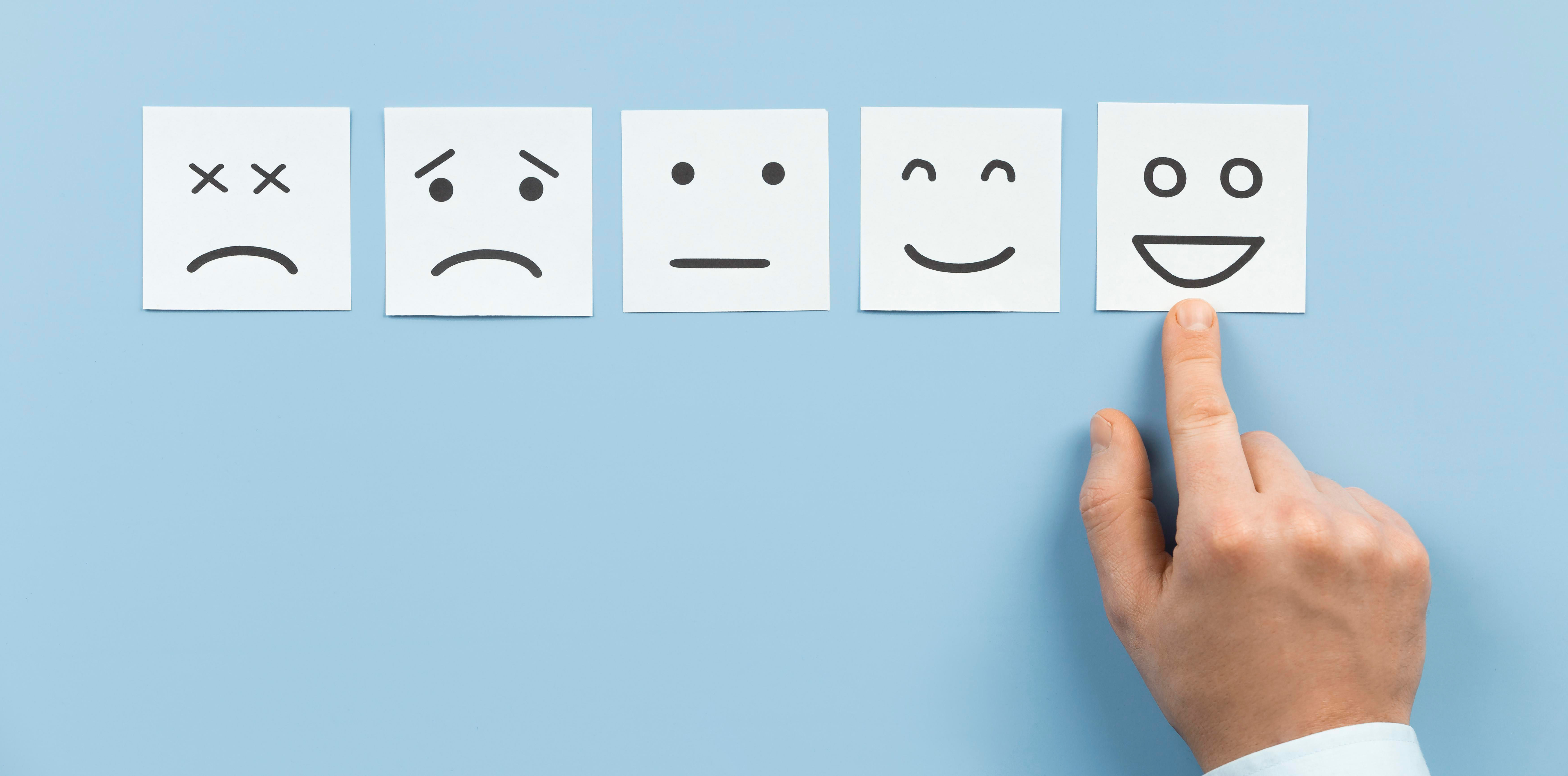 Photo of five faces drawn on white paper squared ranging from unhappy to very happy. A hand is pointing to the happy face. 