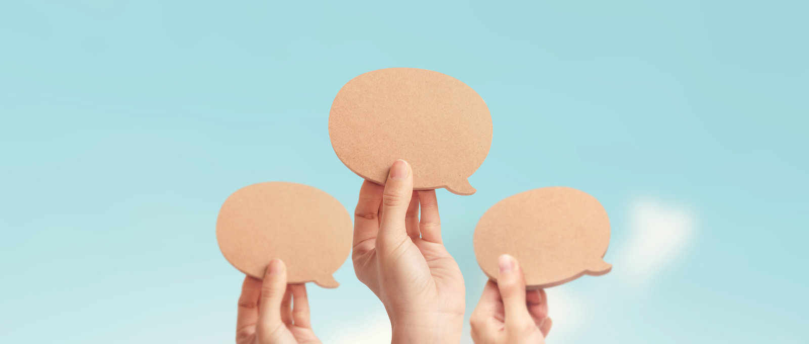 close-up of three hands holding up blank wooden speech bubbles against a pale blue sky
