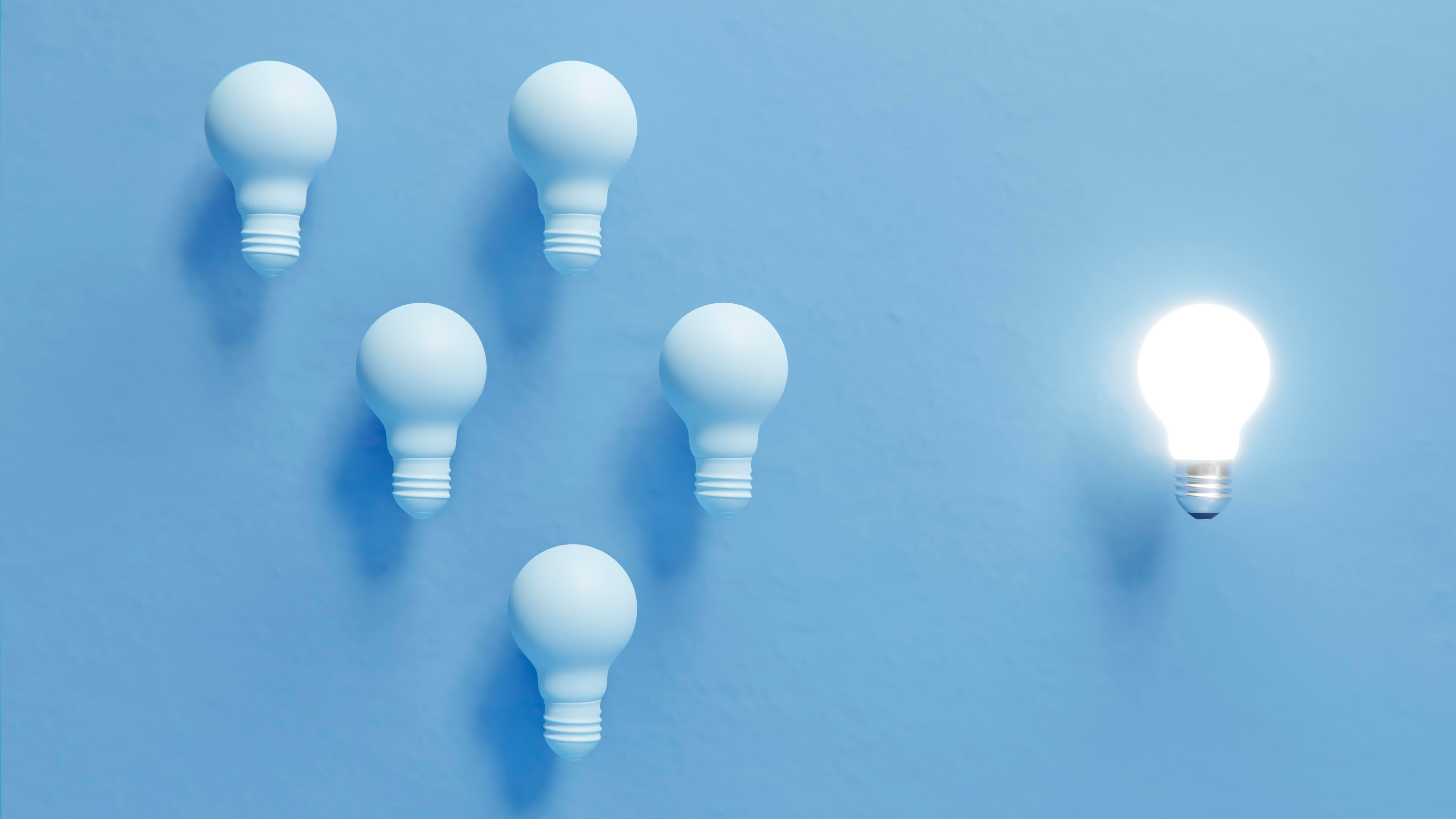 Abstract photo of lightbulbs against a light blue background. Several are dark and grouped together. One is out on its own and lit up.