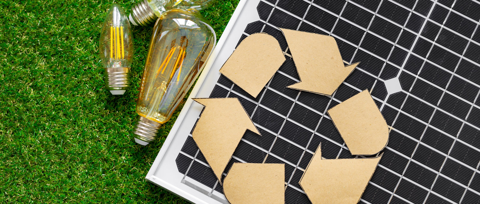 Photo of a solar panel with a paper cut out of the recycling symbol on top of it, together with some low energy lightbulbs, all lying on grass