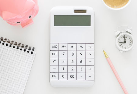 Ariel view of white office desktop with calculator, piggy bank, pen, note pad, diary, calendar and house plant