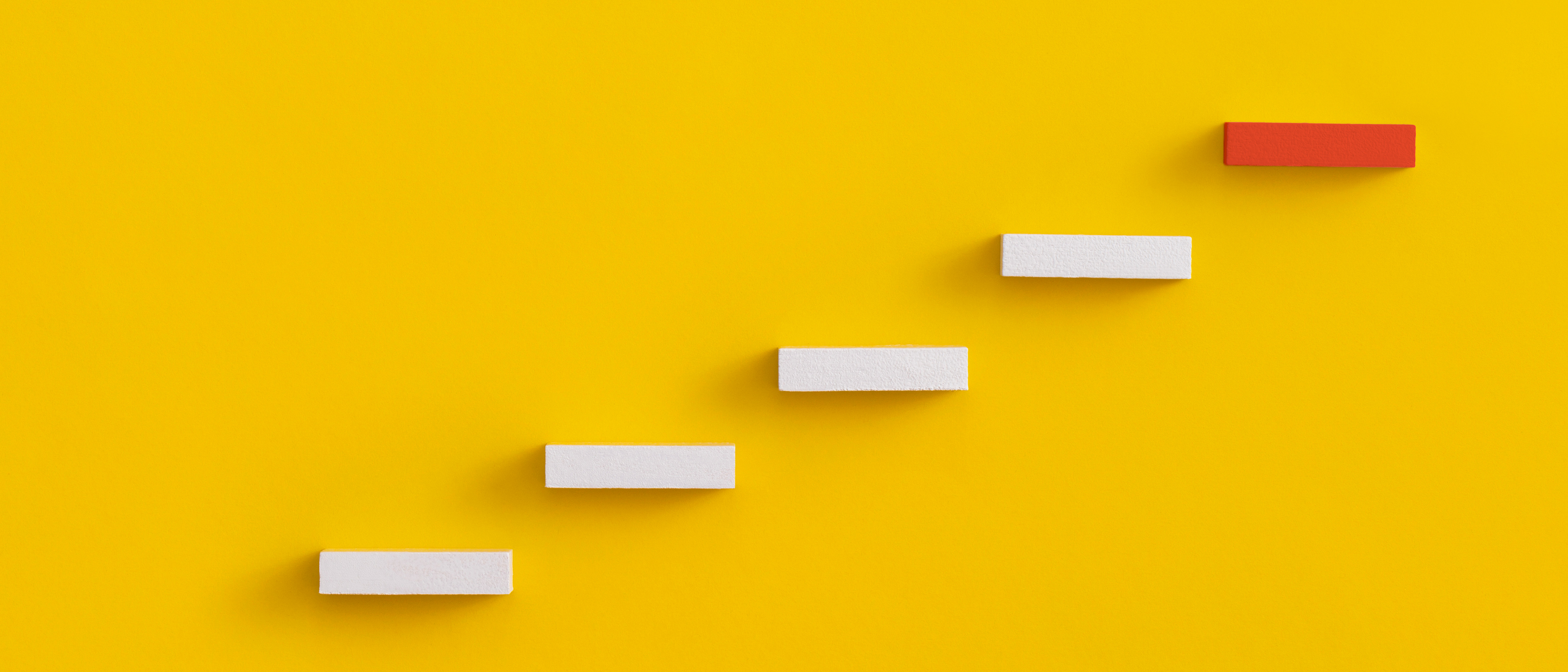 Abstract photo of wooden 'steps' against a yellow background. They go up and right from the bottom left side of the image. The top 'step' is red. 