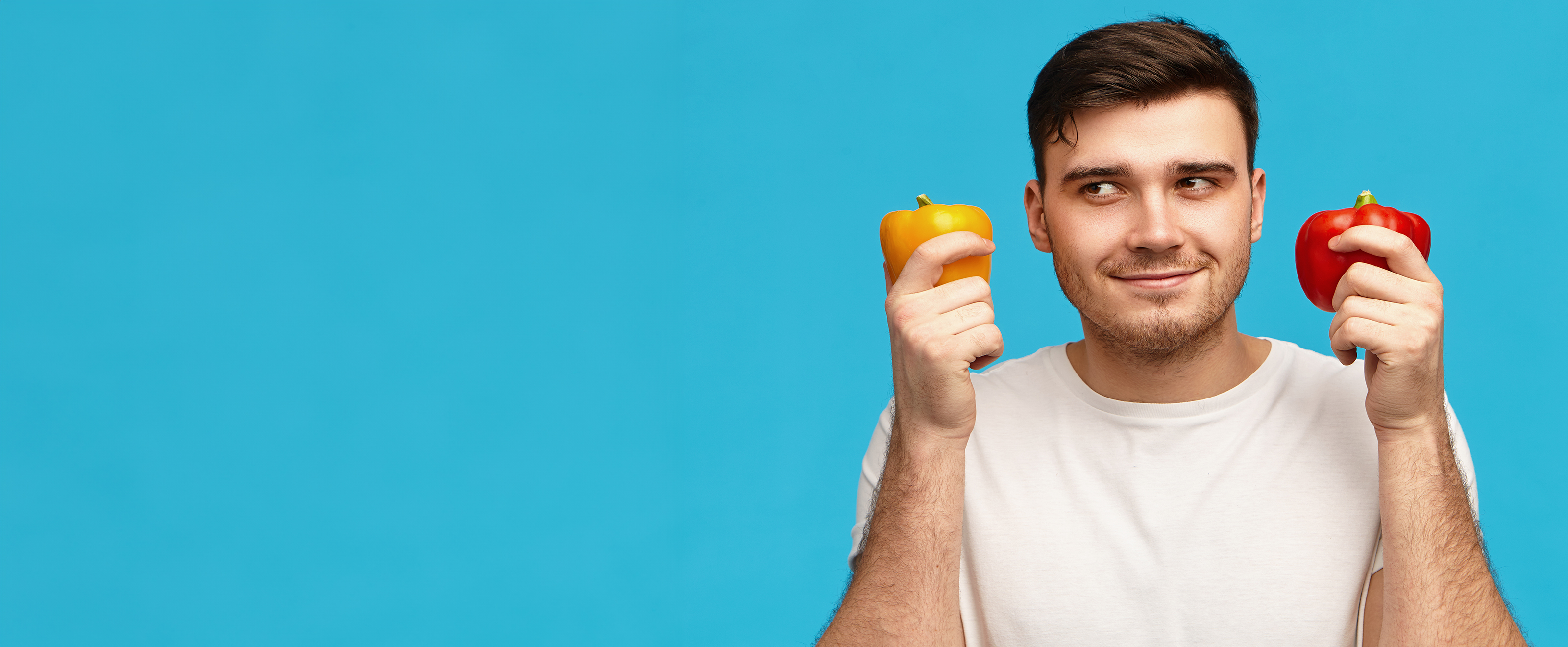 Photo of a man holding a red pepper in one hand and a yellow pepper in the other hand. Standing in front of a blue background